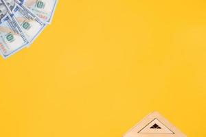 Toy roof of house and one hundred dollar bills on yellow background, flat lay. Concept cost of real estate, rental apartments, investment in construction. Top view, empty space for text photo