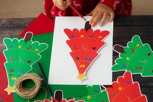 Child draw and make Christmas handmade paper garaland Christmas trees on wooden table. Flat lay. Kid hands. photo