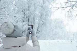 woman in knitted grey hat with pompon from behind taking landscape photo of winter forest snowing with her smartphone