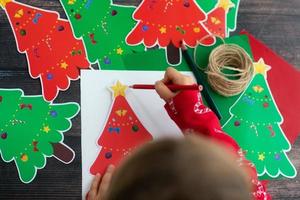 niño dibujar y hacer navidad papel hecho a mano garaland árboles de navidad en mesa de madera. endecha plana manos de niño. foto