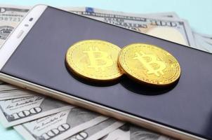 Bitcoins lies on a smartphone and hundred dollar bills on a light blue background photo