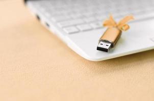 Orange usb flash memory card with a bow lies on a blanket of soft and furry light orange fleece fabric beside to a white laptop. Classic female gift design for a memory card photo