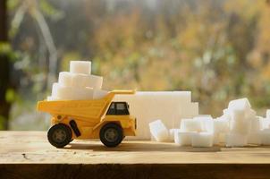 A small yellow toy truck is loaded with white sugar cubes near the sugar debris. A car on a wooden surface against a background of autumn forest. Extraction and transportation of sugar photo