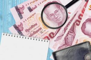Billetes de 100 baht tailandeses y lupa con monedero negro y bloc de notas. concepto de dinero falso. busque diferencias en los detalles de los billetes de dinero para detectar falsos foto