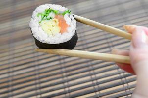 A hand with chopsticks holds a sushi roll on a bamboo straw serwing mat background. Traditional Asian food photo