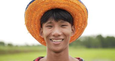 retrato en la cabeza de un agricultor, retrato de un adulto joven con camisa a cuadros y sombrero de pie con los brazos cruzados, riendo y mirando a la cámara con timidez. campo de arroz en el fondo video