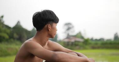 vista lateral y cierre joven granjero sin camisa sentado en el suelo y mirando sus campos de arroz, se siente triste y pensativo video