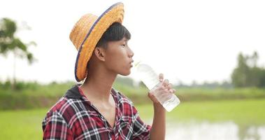 toma en cámara lenta, retrato de un joven granjero con camisa a cuadros y sombrero de paja abierto y bebe agua de una botella para aliviar la sed y el cansancio en el campo de arroz video