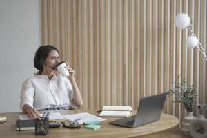 empresaria española positiva satisfecha con auriculares inalámbricos bebiendo café en la oficina