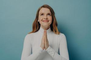 Happy young woman making wish while folded hands in prayer gesture, isolated on blue background photo