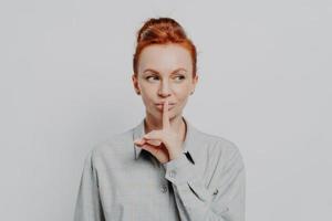 Young ginger woman keeping finger on lips and looking aside while standing against grey wall photo