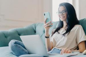 Positive optimistic brunette woman dressed in casual clothes and optical glasses, focused in smartphone display, searches new website for shopping, watches training webinar online, sits on sofa