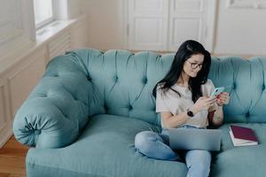 View from above of busy female freelancer sits in cozy room on comfortable sofa, works on new startup project, uses laptop computer, holds mobile phoneand sends text messages, interacts with netbook photo