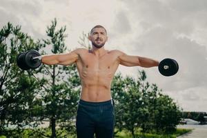 Strong handsome muscular bodybuilder pumps up arm muscles with barbells, does bidybuilding biceps exercises outdoor, poses with naked torso against green trees and sky, makes weight exercise photo