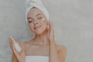 una foto en la cabeza de una mujer atractiva y complacida aplica loción facial, está satisfecha con un nuevo producto cosmético, mantiene los ojos cerrados, toca la piel suave después del baño, tiene una tez bien cuidada, posa contra una pared gris