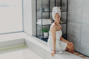 Beauty model with healthy perfect skin applies cream for body care, poses wrapped in towel after taking shower, poses at bathtube, looks thoughtfully away, feels refreshed and relaxed. Spa procedures photo