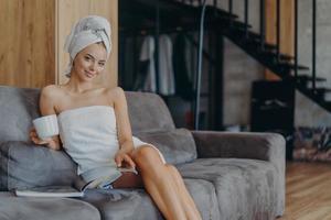 Smiling relaxed young European woman sits on comfortable sofa, reads magazine and drinks coffee, poses on comfortable sofa, undergoes beauty treatments, has satisfied expression. Spending time at home