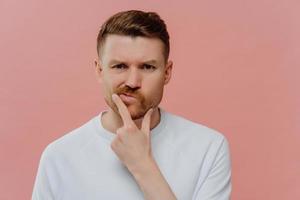 Photo of serious man holds chin purses lips makes decision with doubtful expression focused displeased at camera dressed in casual white t shirt isolated over pink background. Let me thinks a bit