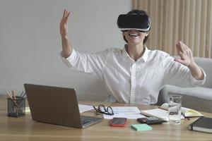 Joyful young business woman moving hands in air while using virtual reality glasses at workplace photo