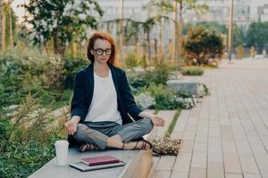 Young relaxed redhead woman meditates in park feels calm sits in lotus pose outdoor photo
