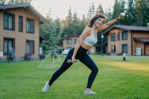 una foto lateral de una mujer decidida y sonriente activa levanta pesas y hace ejercicio matutino, vestida con ropa activa, posa en el césped verde contra casas privadas. deportista entrena bíceps con pesas