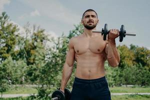 Muscular shirtless young man raises barbells outdoors, trains muscles and has strong body. Athletic sportsman with strong arms leads healthy lifestyle, enjoys training against nature background. photo