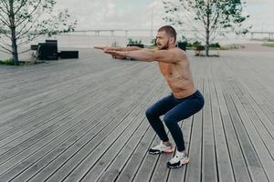 Full length shot of active man does squatting exercise poses with naked torso has muscular body poses outside. Strong athletic fit man in sportswear exercises in open air. Confident fitness instructor photo