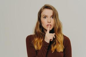 Mysterious caucasian teen girl holding finger on lips, looking at camera with scared face expression photo