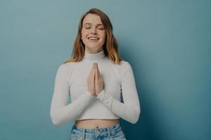 Smiling young woman holding hands folded in prayer gesture, keeping eyes closed in hope photo