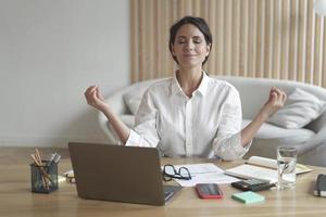 Attractive female entrepreneur sits in relaxed position at desk in front of laptop with eyes closed photo