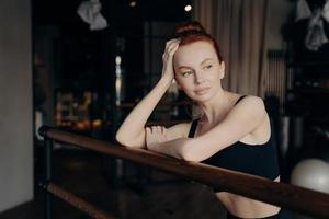 Young attractive woman with red hair stands leaning on ballet barre and resting after workout photo