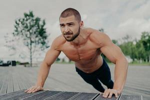 Active muscular man does push up exercise poses shirtless outdoor, exercises in park, has thick beard, stands in lower position. Motivated bodybuilder does workout regularly. Athlete trains upper body photo