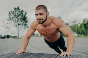 el hombre musculoso activo empuja hacia arriba el ejercicio posa sin camisa al aire libre, hace ejercicio en el parque, tiene barba gruesa, se para en una posición más baja. el culturista motivado hace ejercicio regularmente. atleta entrena la parte superior del cuerpo foto