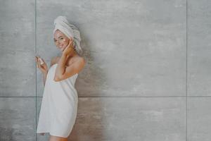 mujer joven feliz y saludable con piel facial tierna, aplica crema de belleza y se somete a tratamientos de belleza, envuelta en toalla de baño, posa contra fondo gris con espacio para su promoción