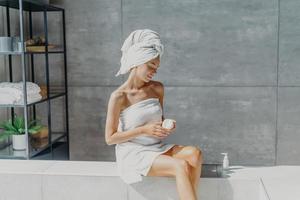 Relaxed young European woman applies moisturizer cream on legs after taking bath sits wrapped in towel in bathroom, enjoys beauty treatments, uses cosmetic product for healthy skin. Hygiene concept