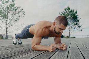 Outdoor shot of strong sportsman stands in plank pose, puts all efforts in keeping fit and healthy. Athlete man with concentrated expression, works out in open air. Bodybuilder makes press exercise photo