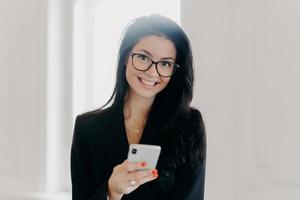 Happy business consultant dressed in formal wear, holds mobile phone, sends text messages, reads news online, poses indoor at office, has hard working day. People, technology, entrepreneurship concept photo