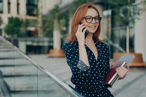 Positive young redhead woman enjoys mobile phoning keeps smartphone near ear photo