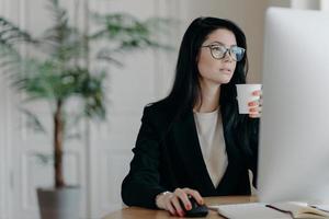 Busy young female administrative poses at desktop, drinks hot coffee from paper cup, dressed formally, develops business strategy, plans startup, involved in working process. Occupation concept photo