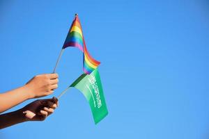 Saudi Arabia flag and rainbow flag, LGBT symbol, holding in hands, blue sky background, concept for LGBT celebration in Saudi Arabia in pride month, June, soft and selective focus.