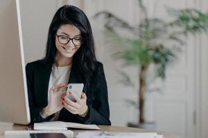 Beautiful smiling female freelancer works remotely, concentrated in smartphone, sends feedback on received message, sits in coworking space, dressed formally, works on computer, surfs internet photo