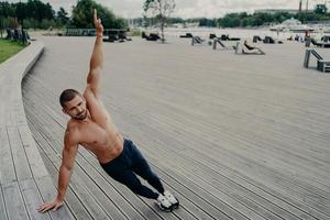 Self determined muscular man dressed in sport clothes stands in side plank, keeps arm raised, has workout outdoor during summer day. Sport, healthy lifestyle, strength and endurance concept. photo