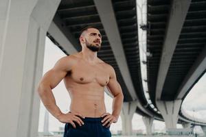 Athletic male fitness model keeps hands on waist poses with muscular perfect naked body, looks seriously into distance, does sport exercises outdoor, focused somewhere, goes in for sport regularly photo