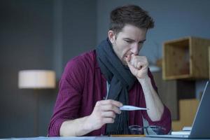 Unhealthy looking male entrepreneur in scarf around his neck coughing looks at digital thermometer photo