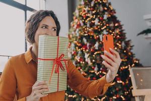 alegre morena brillante española haciendo selfie en smartphone mientras posa con regalo de navidad foto