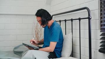 Young Asian man using tablet checking social media feeling happy smiling while lying on bed after wake up in the morning, Attractive male smiling relax in bedroom at home concept. photo