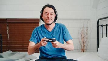 Asian man using joystick playing video games in television in bedroom, male feeling happy using relax time lying on bed at home. Men play games relax at home concept. photo