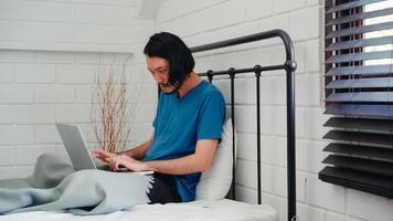 Young Asian man using laptop checking social media feeling happy smiling while lying on bed after wake up in the morning, Attractive male smiling relax in bedroom at home concept. photo