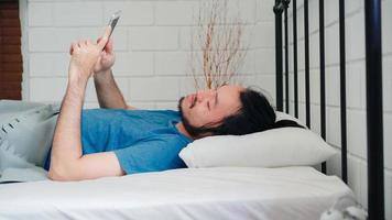 un joven asiático se despierta por la mañana, un hombre asiático que usa un teléfono móvil revisa las redes sociales y envía un mensaje después de despertarse en la cama en el dormitorio de su casa. los hombres guapos toman una siesta, se relajan con sueño en el concepto de casa moderna. foto