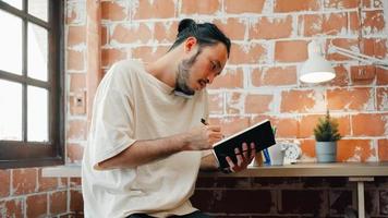 Freelance Asia guy working at his desk in office taking a call on his mobile phone while writing notes on a notepad. Working from home, remotely work, social distancing, quarantine for coronavirus. photo